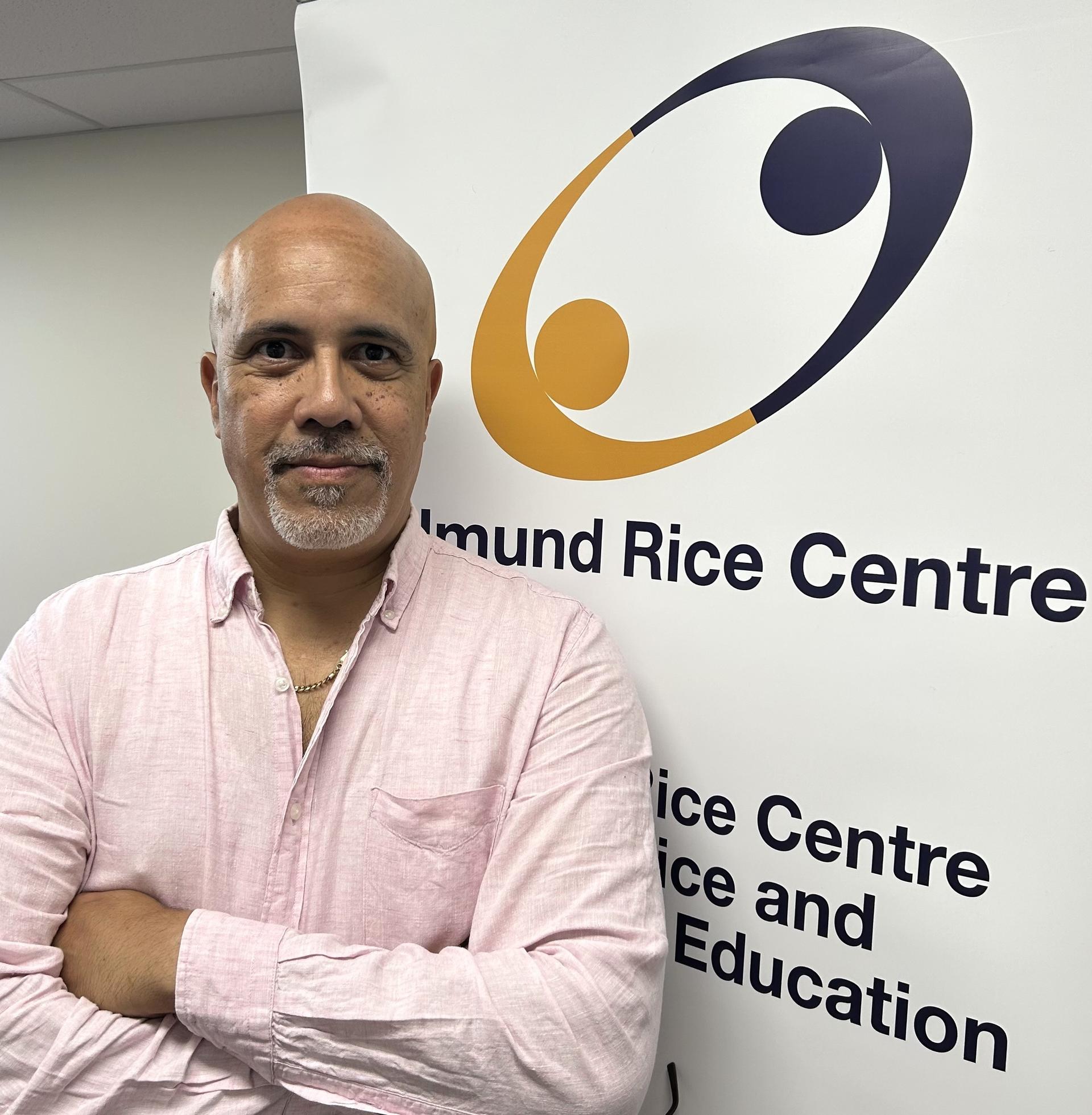 A man in a pink shirt stands confidently beside a white banner with a logo and text.