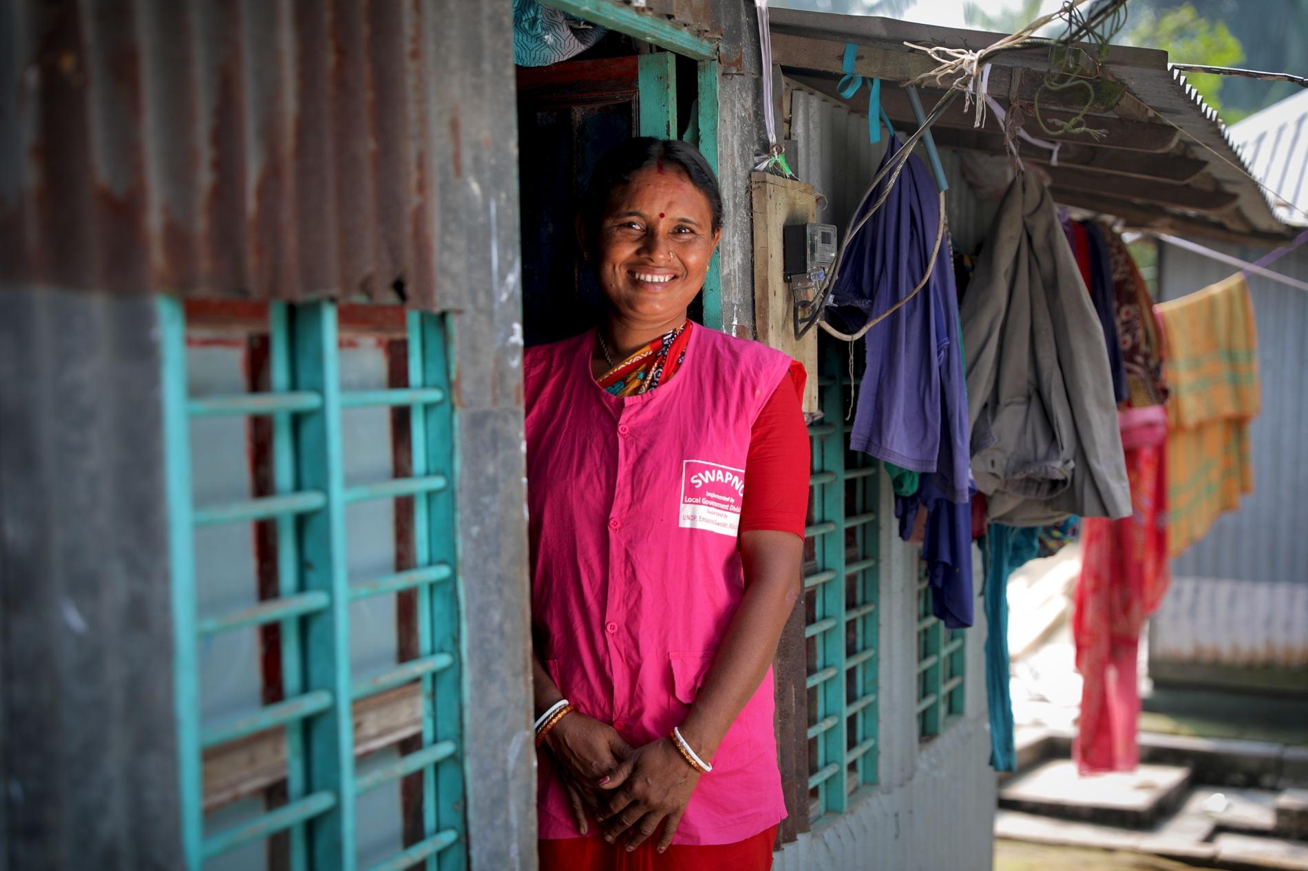 Smiling woman in a pink vest stands in doorway of a colorful house with clothes hanging nearby.