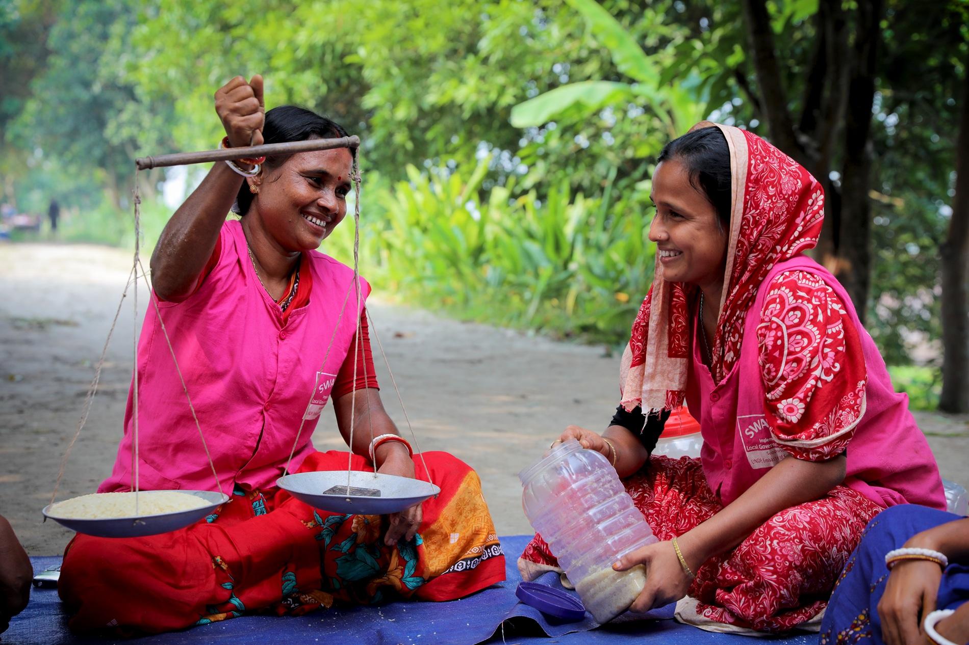 Two women in bright clothing sit outdoors, smiling while weighing ingredients on a scale.