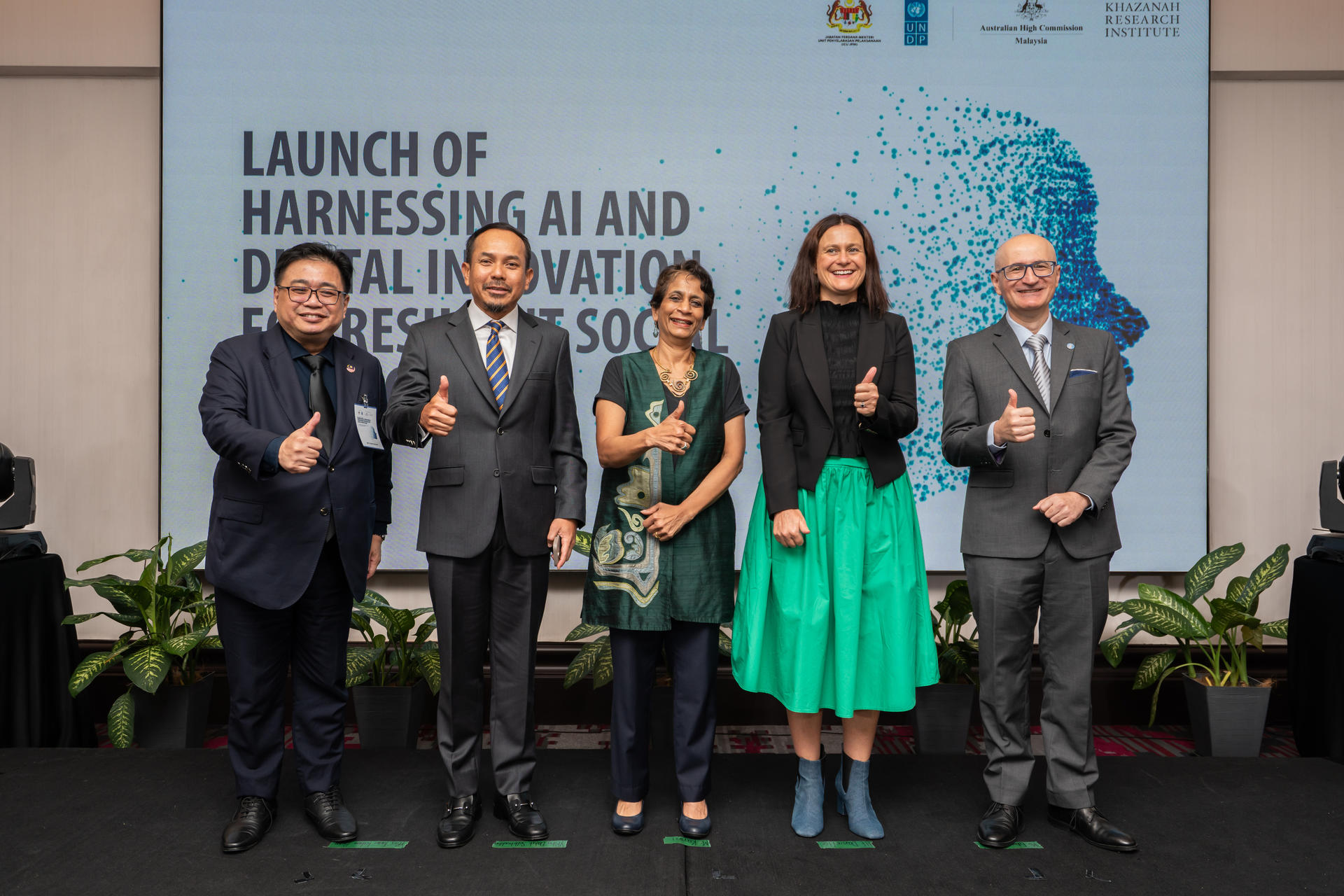 Five people standing together, smiling and giving thumbs up, in front of a presentation backdrop.
