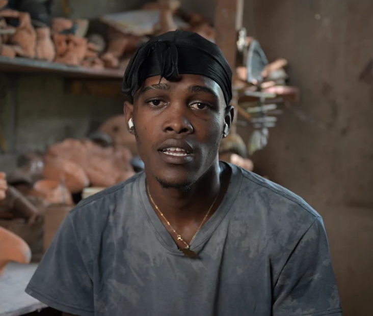 A young man in a gray shirt speaks in a workshop filled with clay sculptures.