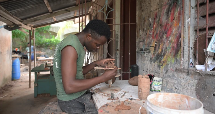 A focused young man sculpting clay at a workbench in a rustic workshop.