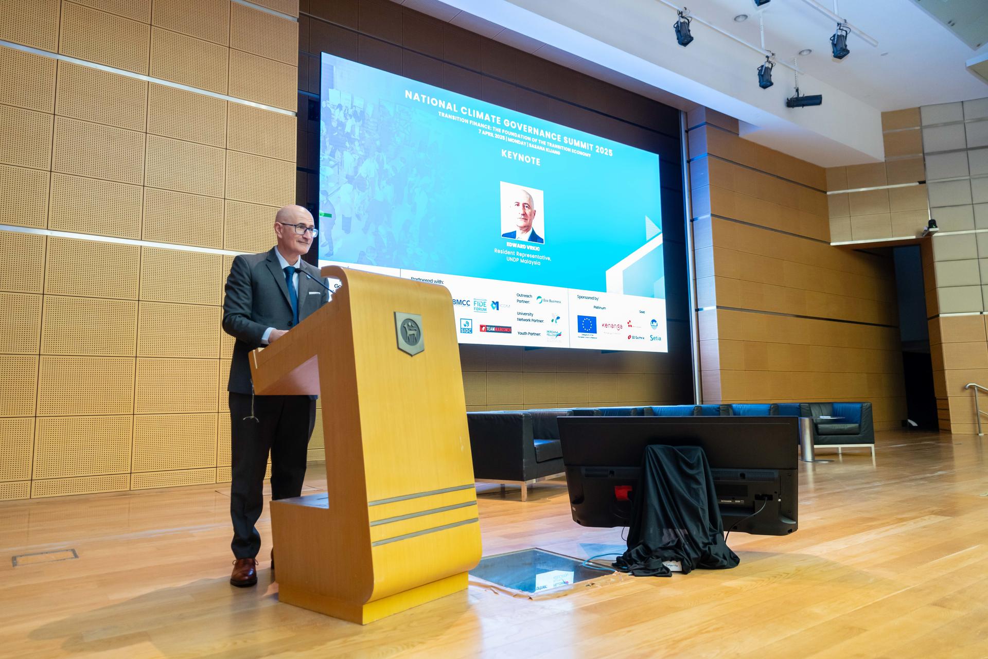 A speaker stands at a podium in a seminar room, facing a large presentation screen.