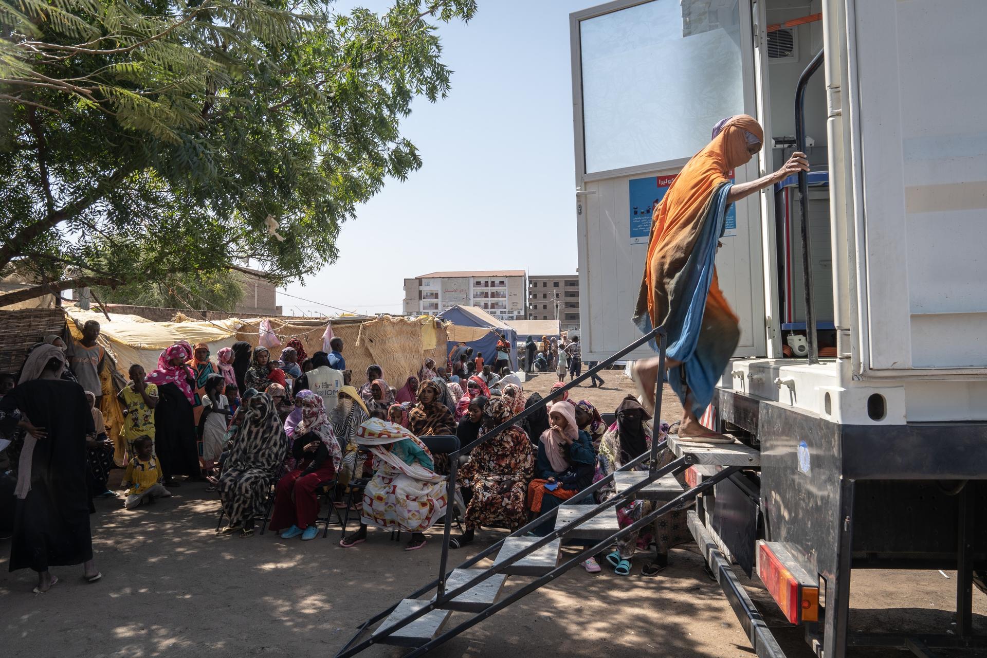 A person in traditional clothing climbs stairs into a medical unit, with a crowd waiting nearby.