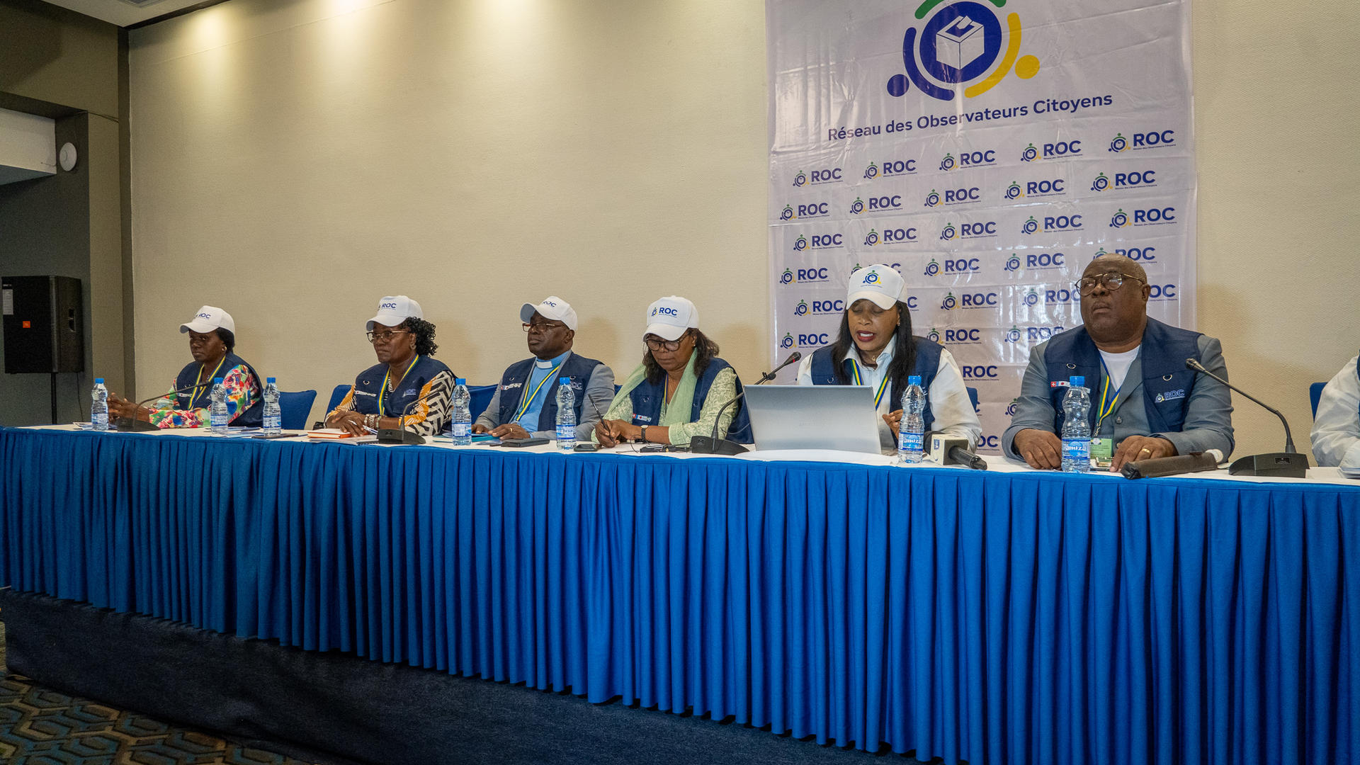 A group of six people seated at a blue table, with a banner behind them, in a conference setting.
