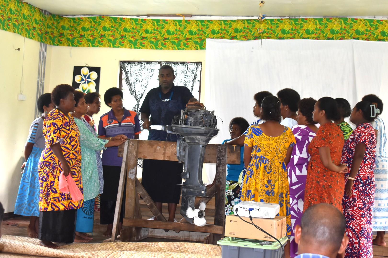 A group of women in colorful attire attentively watches a man demonstrate an engine.