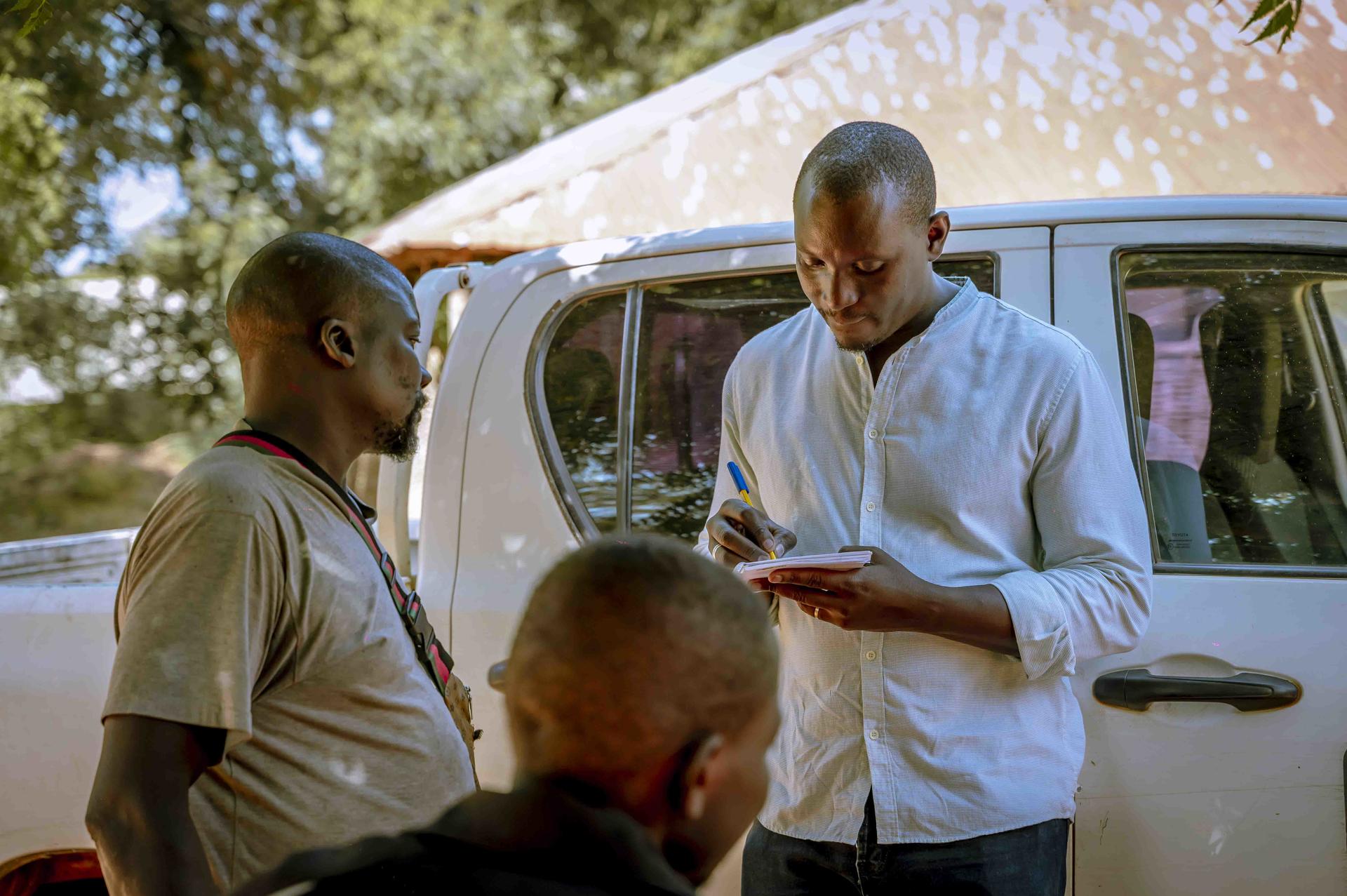 A man in a white shirt takes notes while talking to two men outdoors near a vehicle.