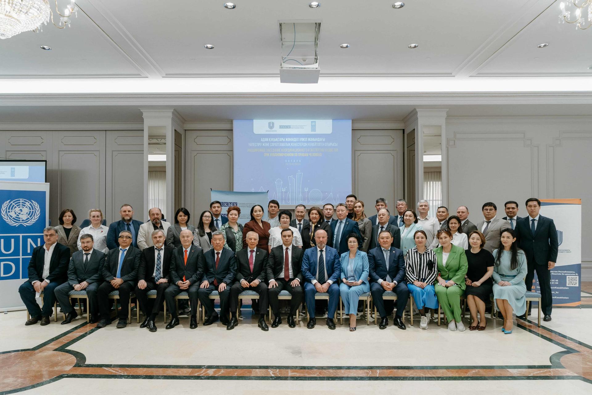 A diverse group of around 40 people poses for a formal group photo in a conference setting.