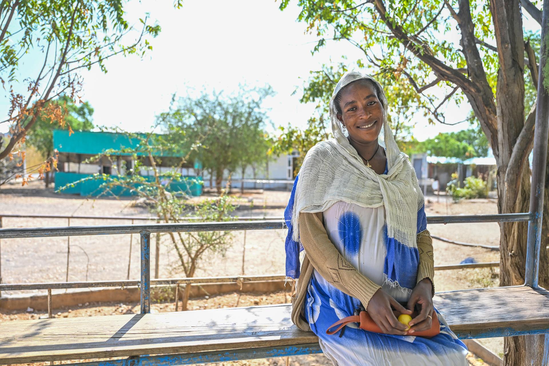 A woman in traditional attire sits outdoors, smiling, with trees and buildings in the background.