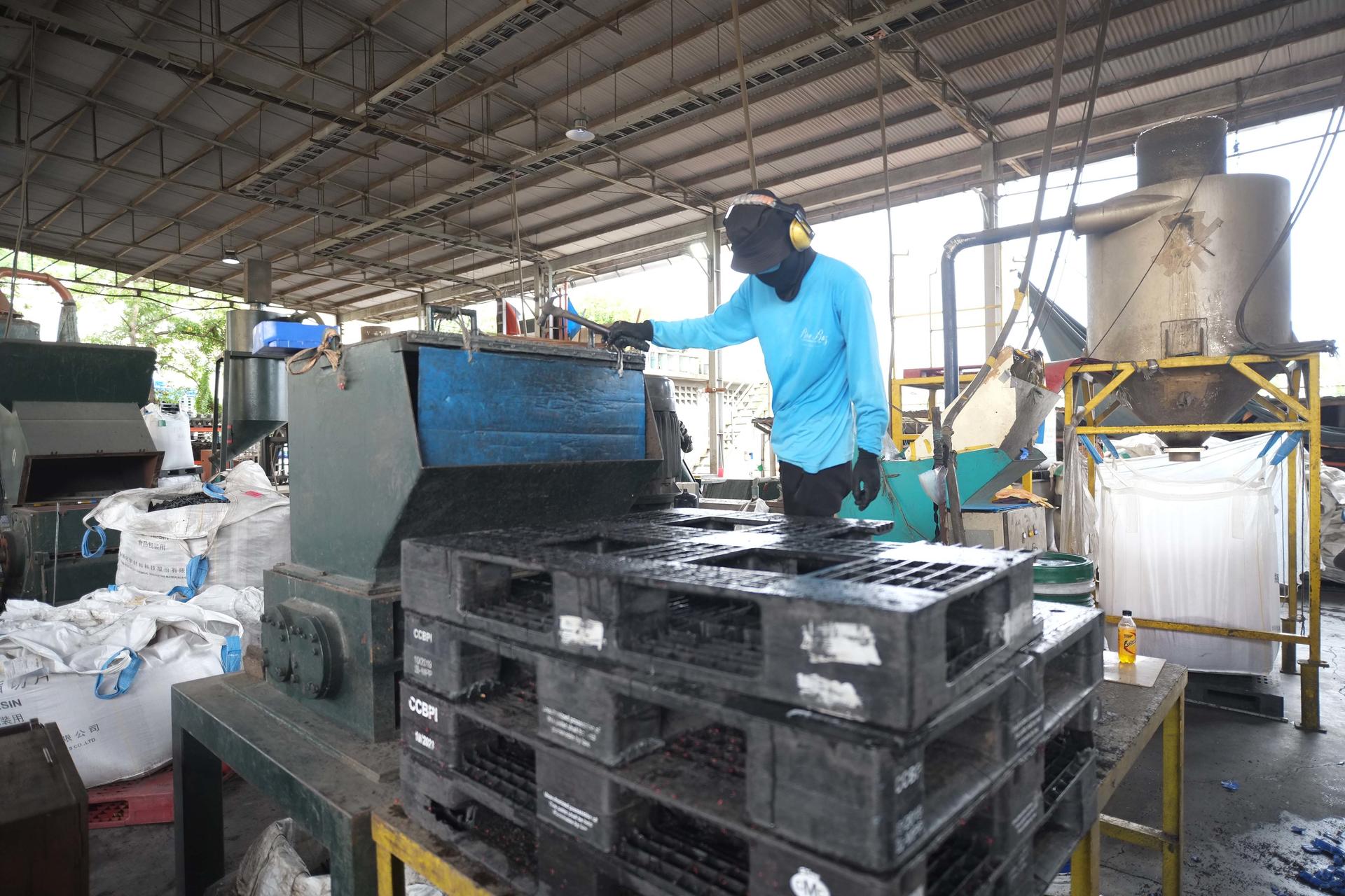 A worker in a blue shirt operates machinery in a factory, with plastic pallets stacked nearby.
