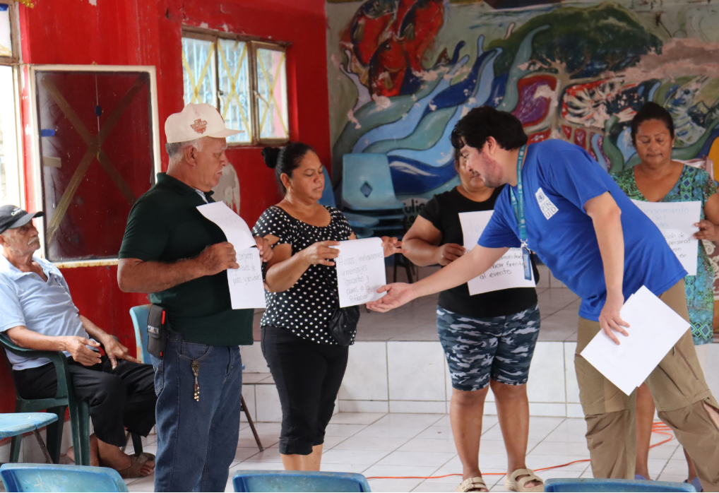 A group of people in a colorful room, exchanging documents during a meeting.