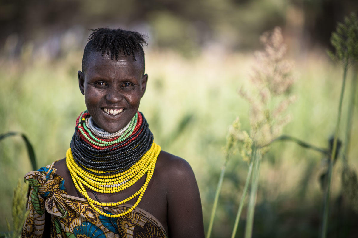 Smiling young woman in colorful beaded necklaces stands amidst tall grass.
