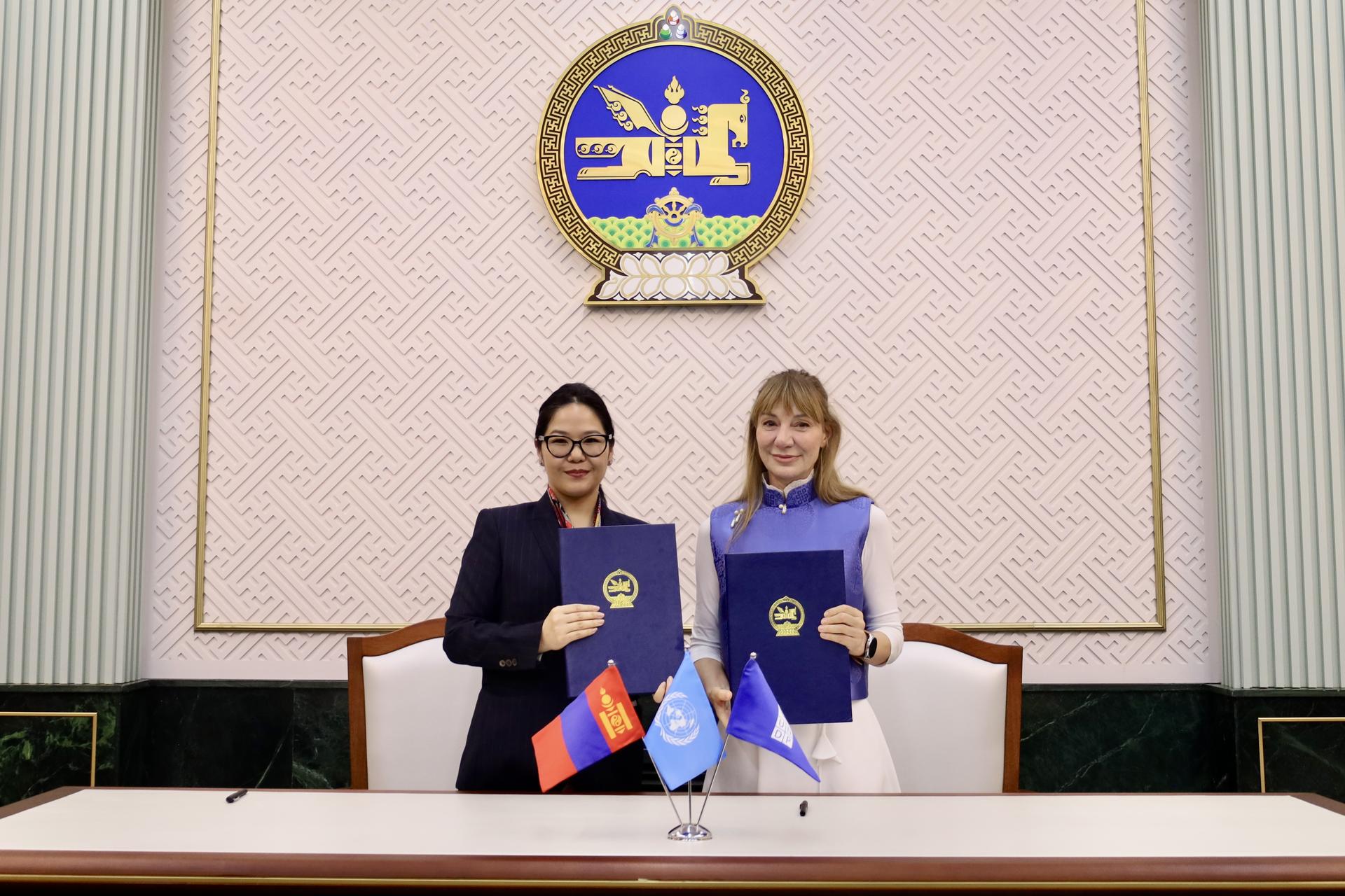 Two women holding documents, smiling at a table, with flags and a logo in the background.