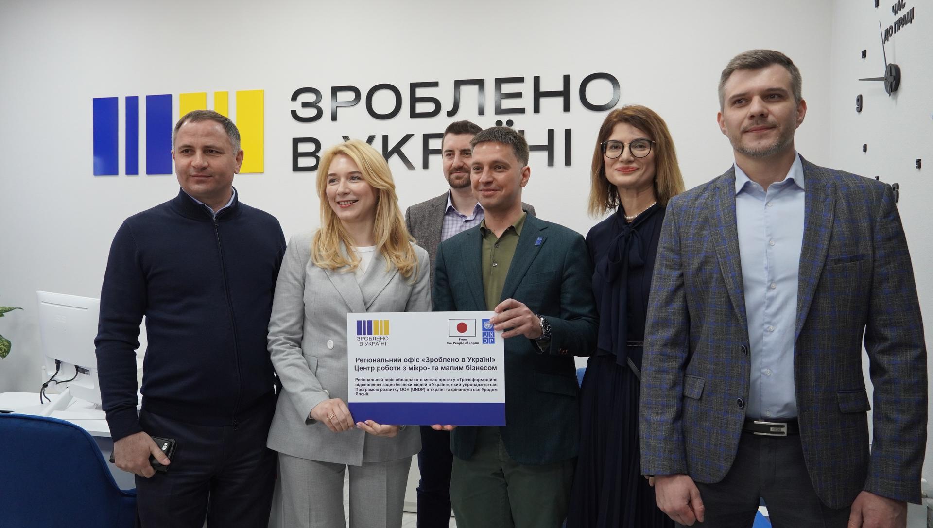 A group of five professionals stands in an office, holding a sign, smiling.