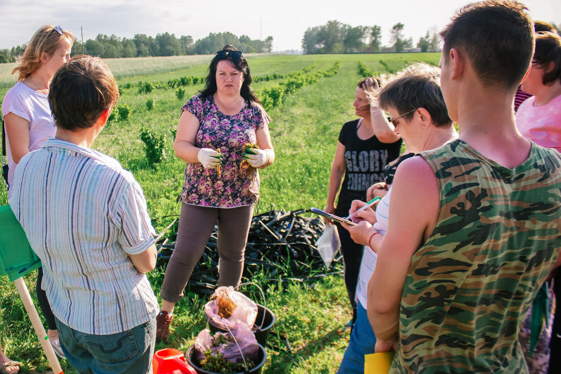A group of people listen to a woman speaking in a green field, surrounded by plants.