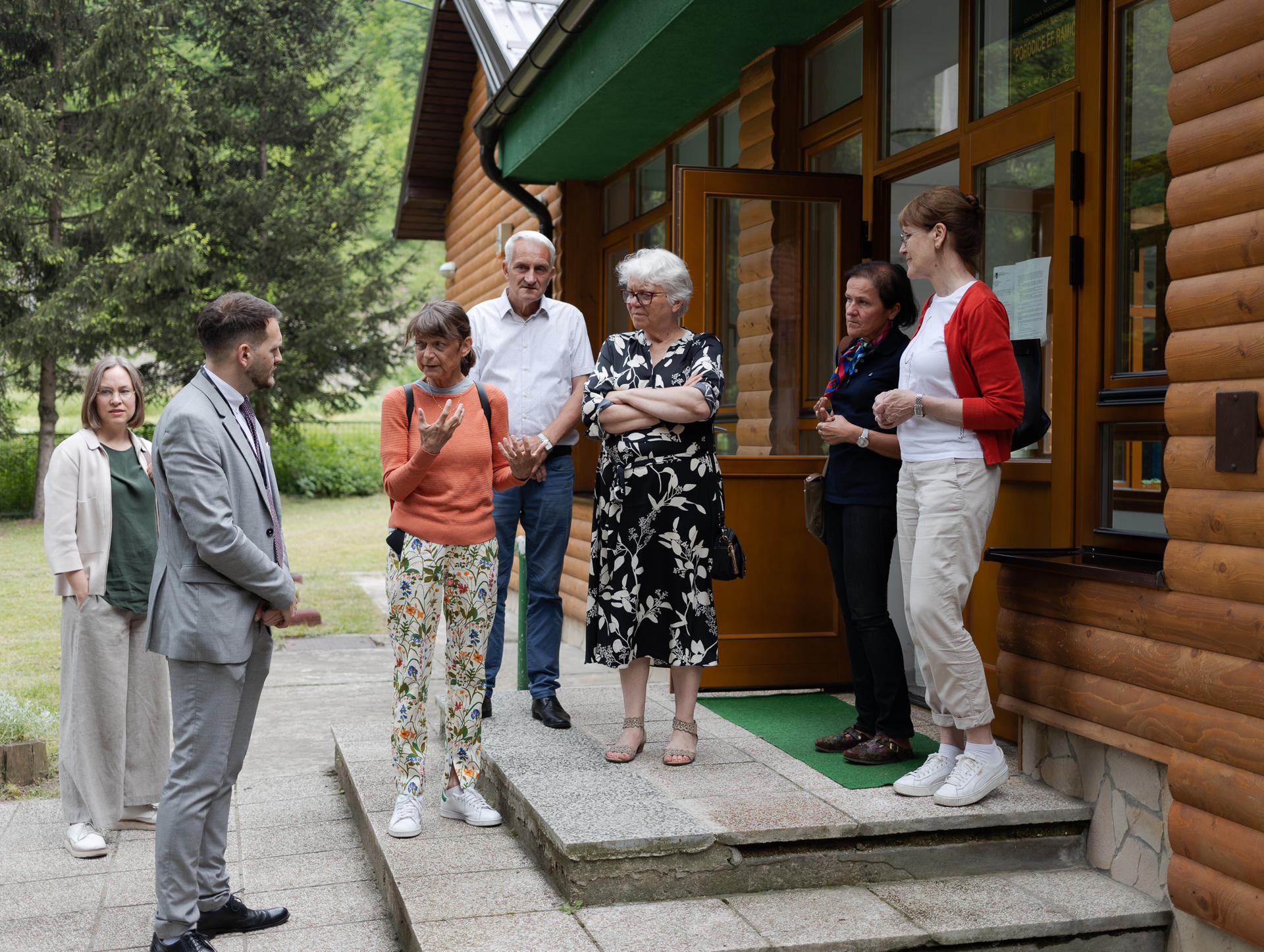 A group of seven people stands on steps outside a wooden building, engaged in conversation.
