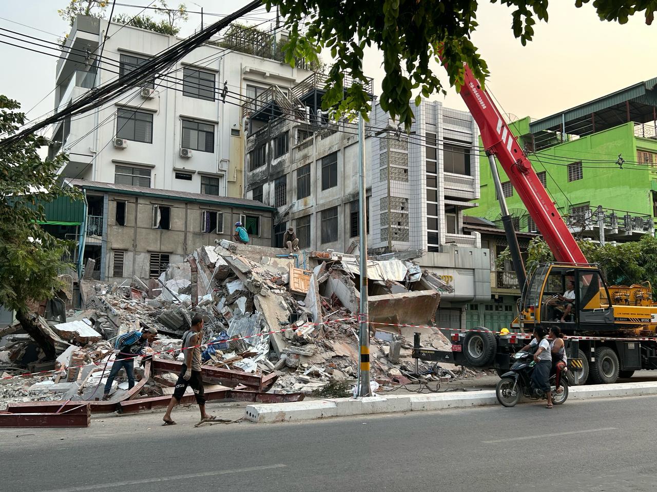 Collapsed building debris with a crane and workers in an urban setting.