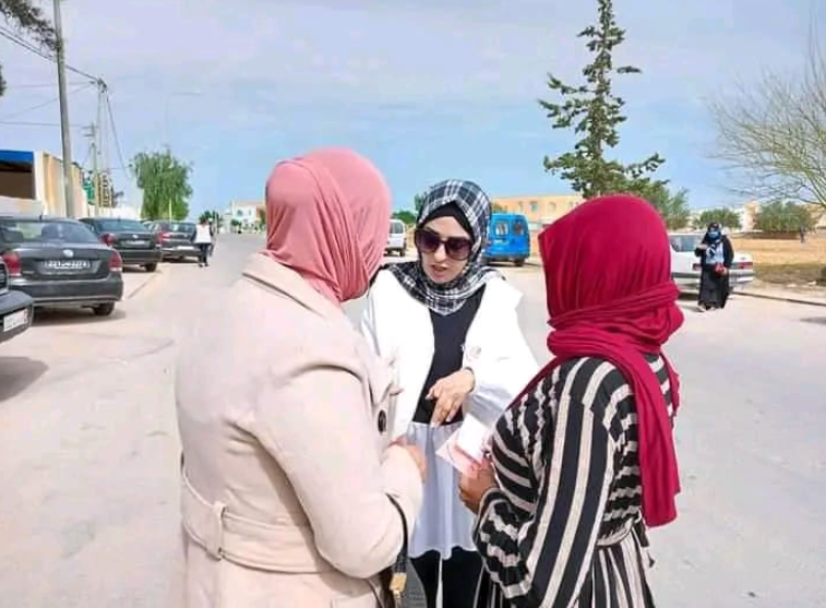 Three women engaged in conversation outdoors, with cars and trees in the background.