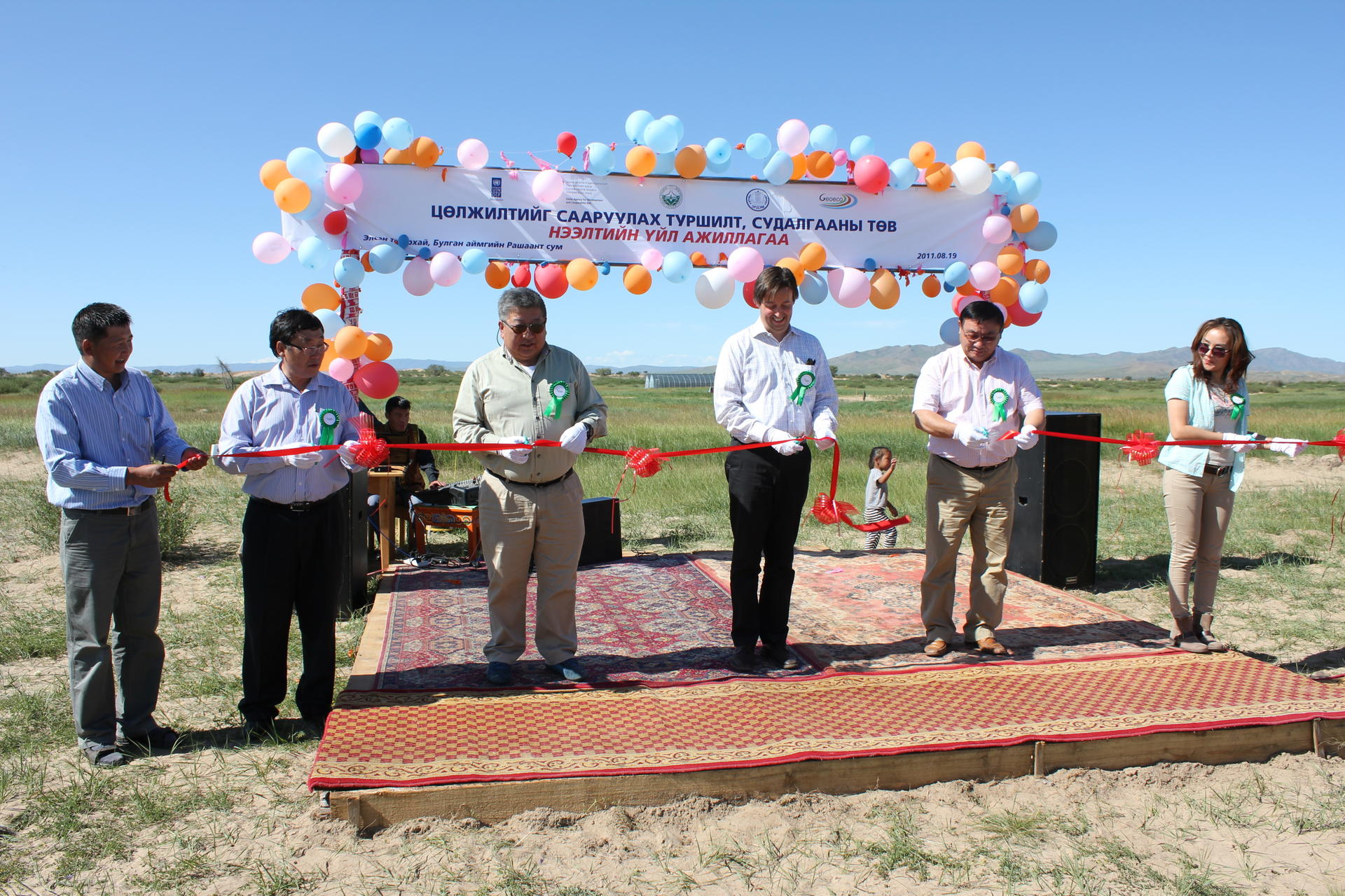 Five individuals at a ribbon-cutting ceremony with balloons and decorations in an outdoor setting.