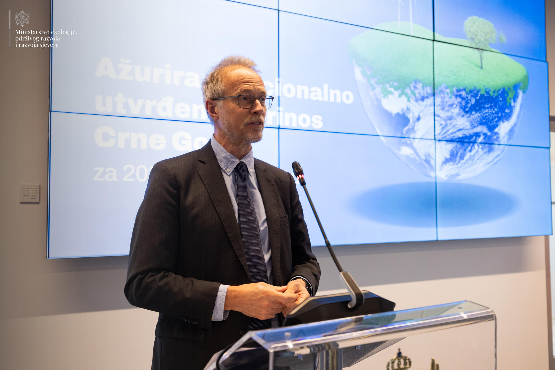 A man in a suit stands at a podium, speaking in front of a large digital screen.