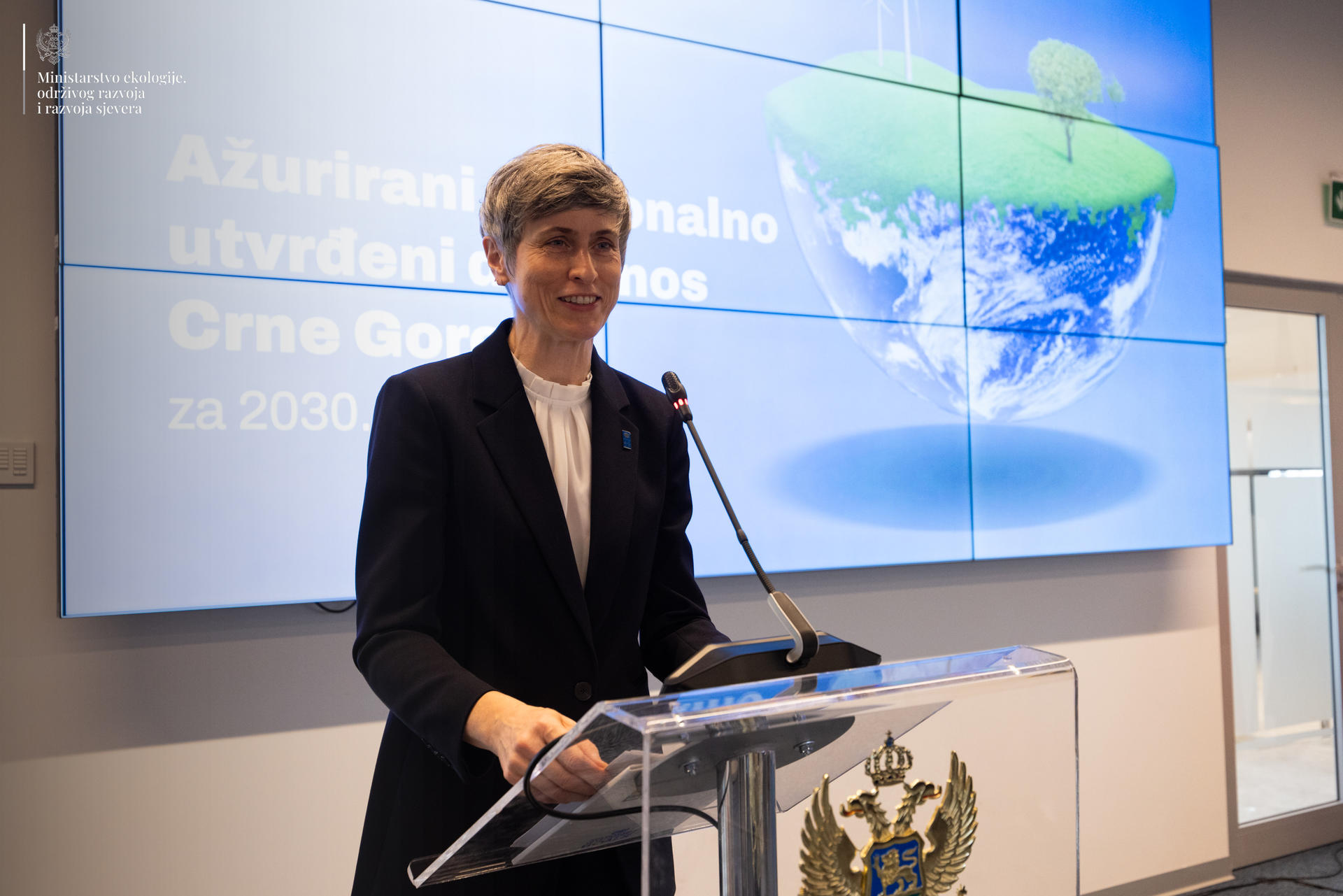 A woman stands behind a podium, smiling, with a global sustainability graphic displayed behind her.