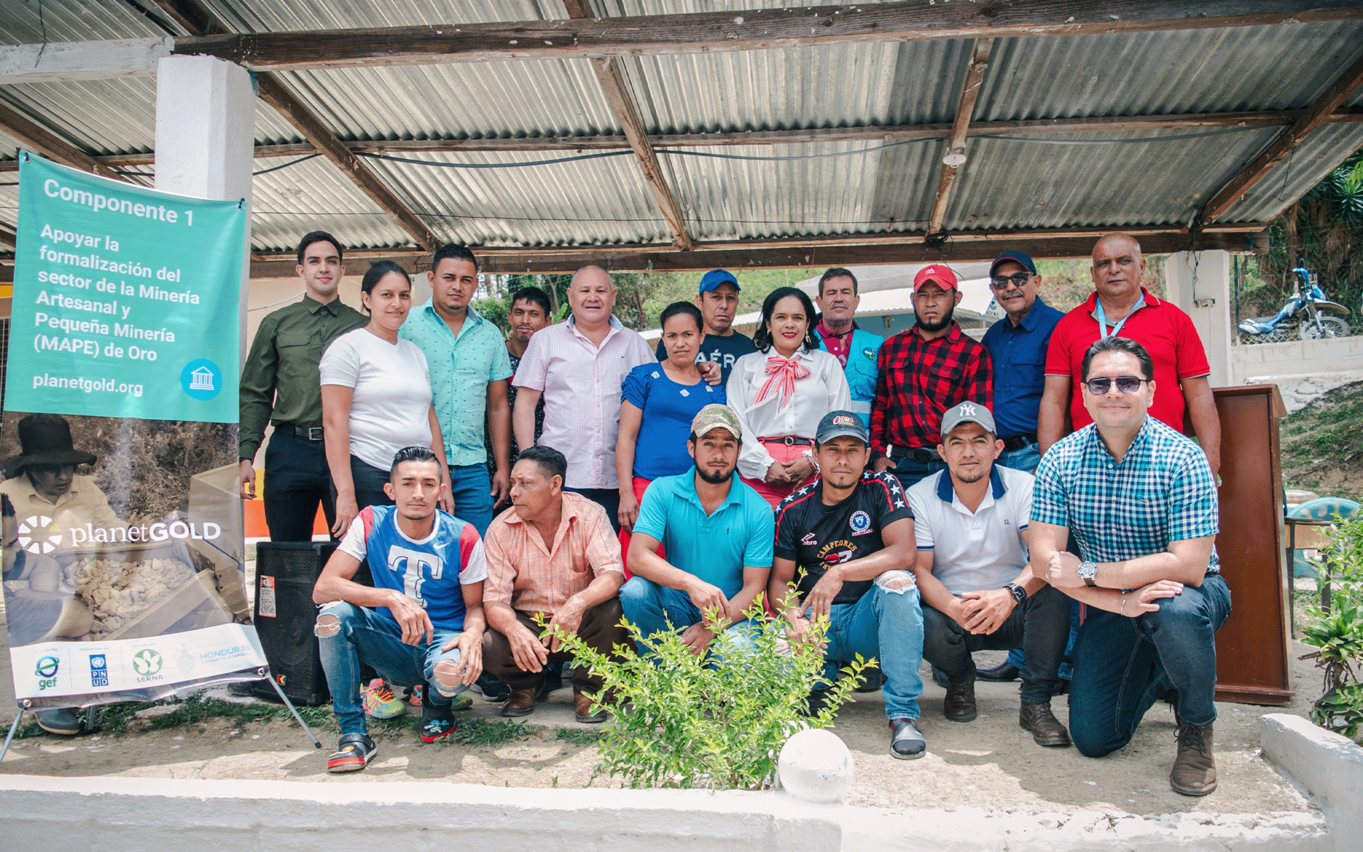 A diverse group of individuals poses together outdoors under a metal roof structure.