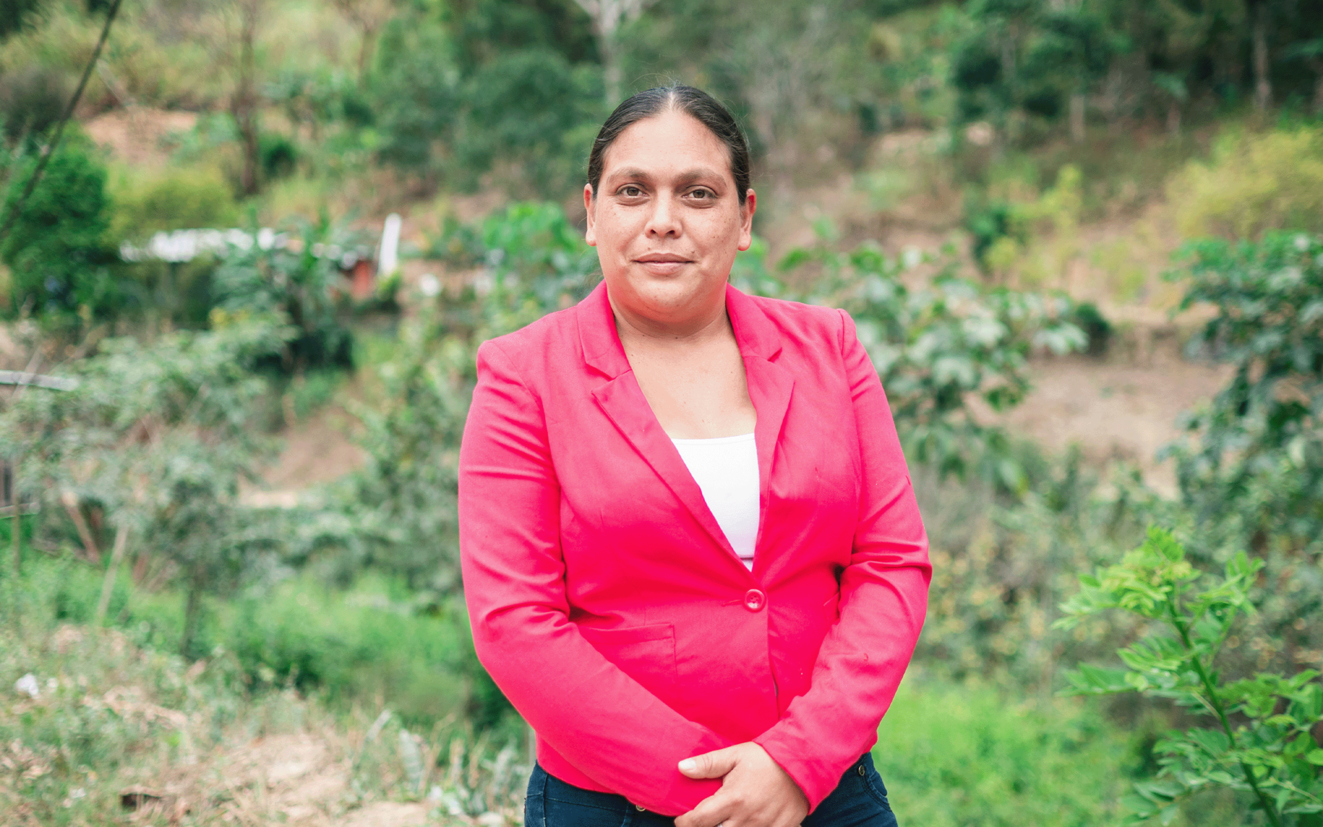 A woman in a pink blazer stands confidently outdoors, surrounded by greenery.