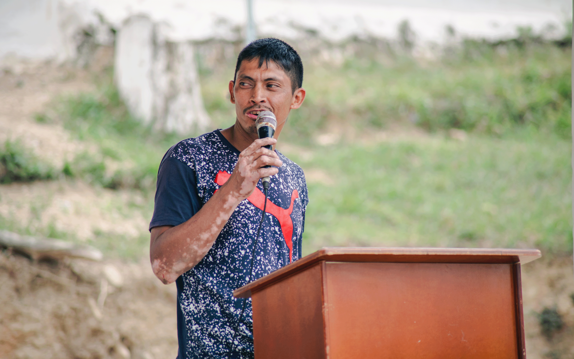 A man speaking into a microphone at a podium outdoors, with green grass in the background.