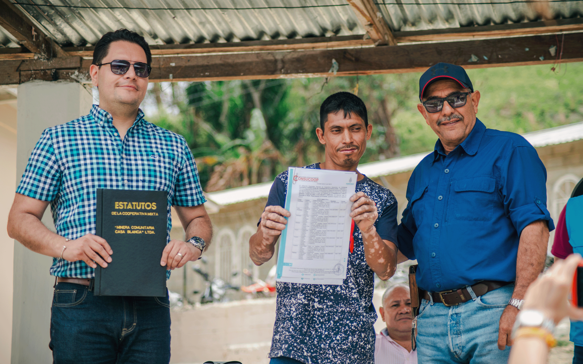 Three men stand under a shelter, smiling while holding a document. Bright greenery in background.