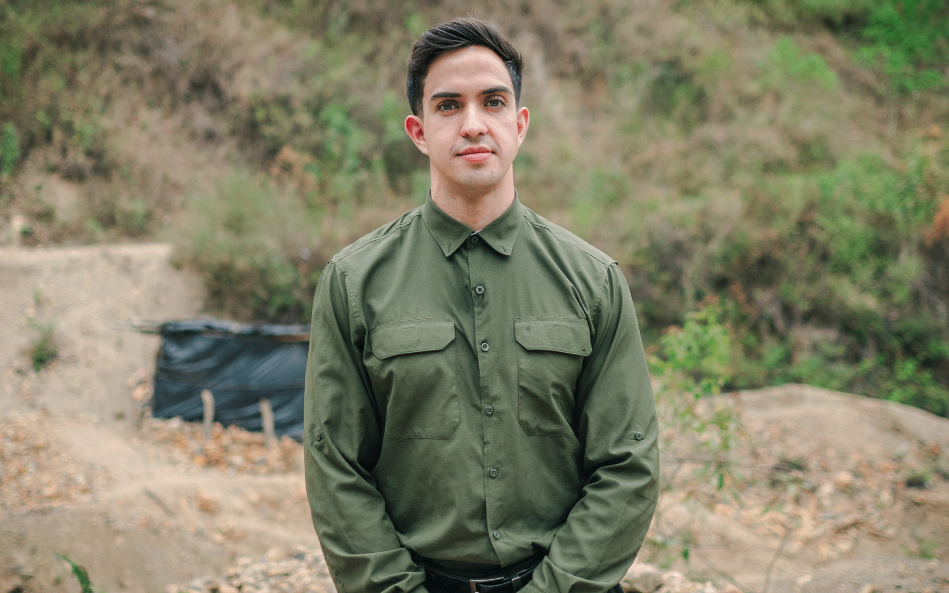 A young man in a green uniform stands confidently outdoors with a hilly background.
