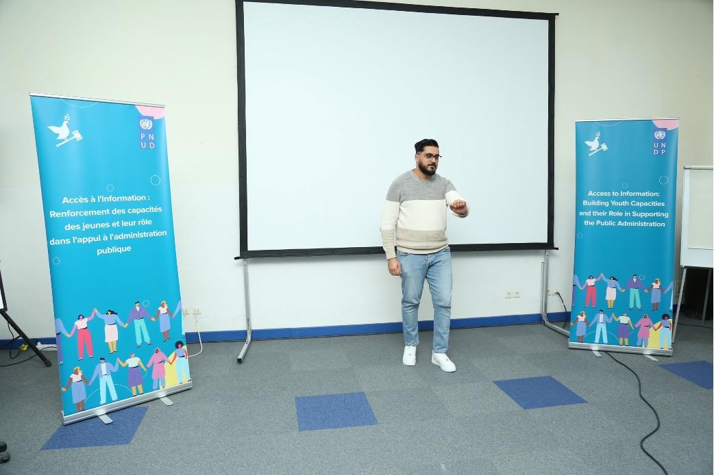A man stands on stage in front of a blank screen, flanked by promotional banners.