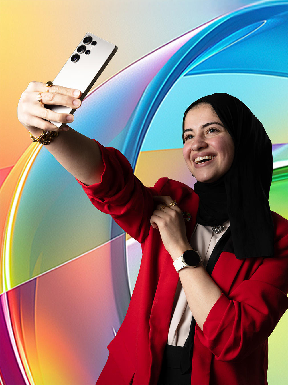 A smiling woman in a red jacket and black hijab takes a selfie against a colorful background.
