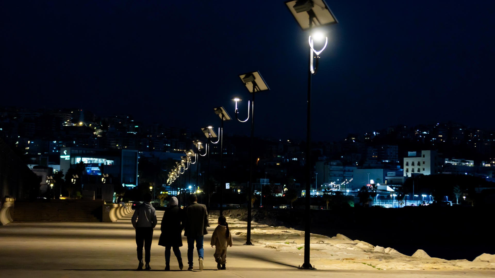 People walking along a lit path at night with city lights in the background.