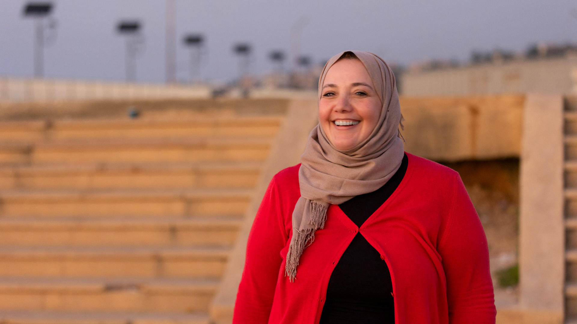 Smiling woman in a red cardigan and scarf stands near stone steps at sunset.