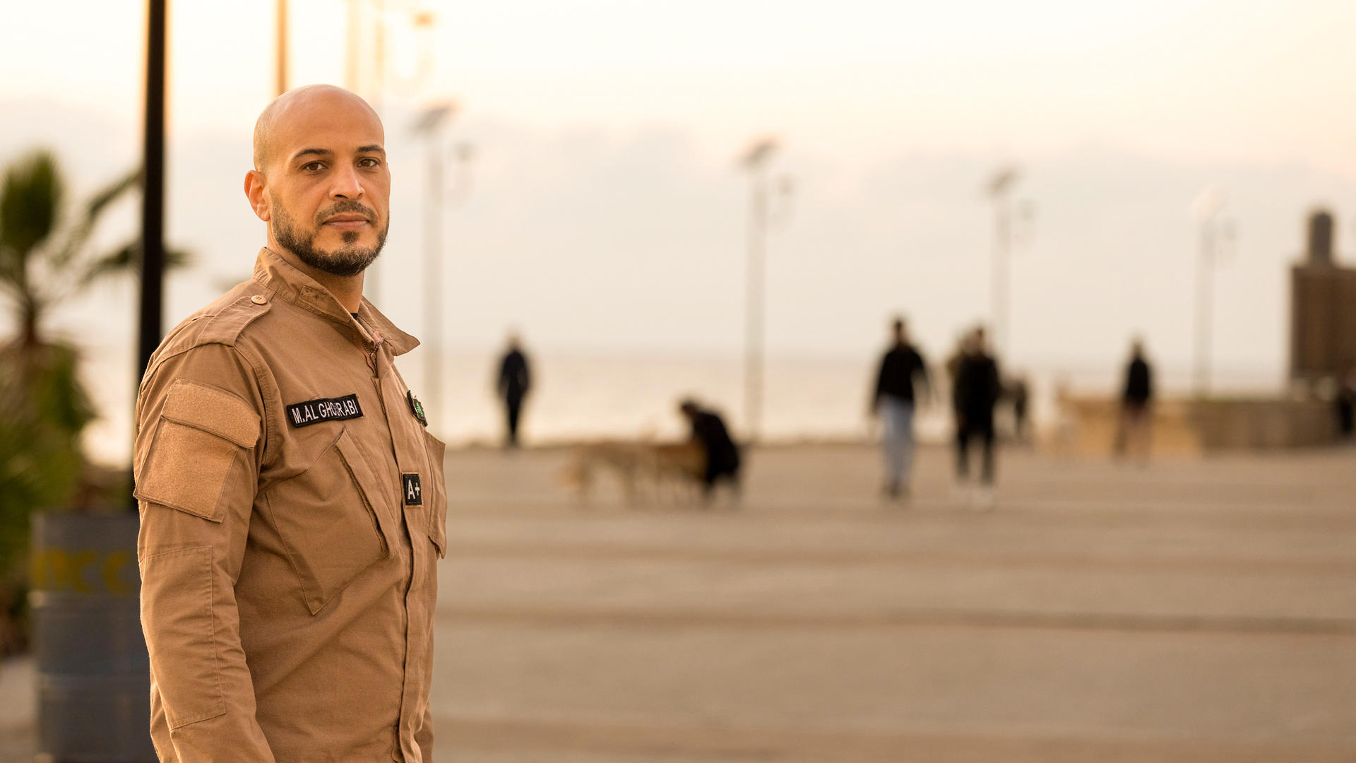 A man in a tan uniform stands on a boardwalk at sunset, people strolling behind him.