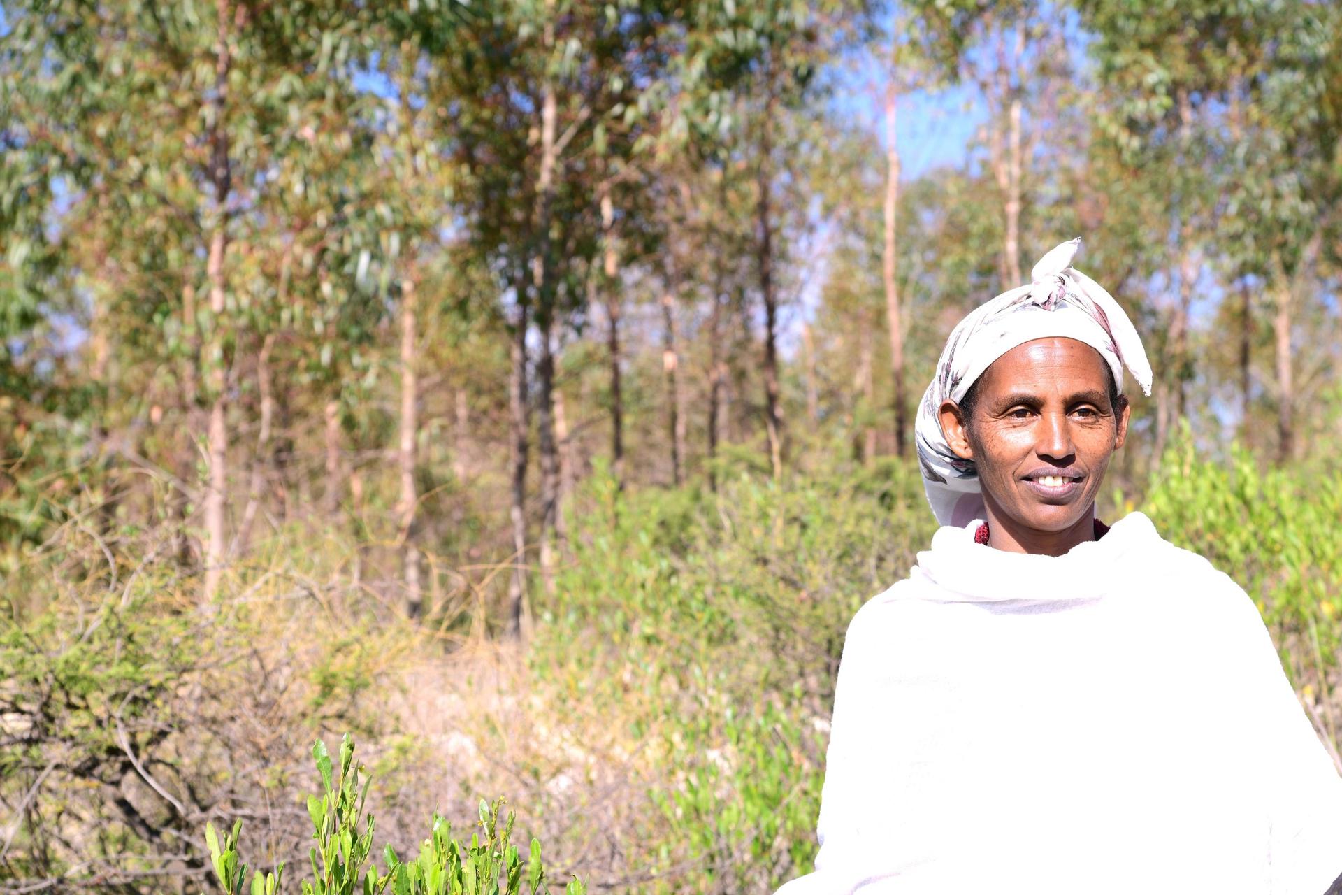 A person in a white garment stands in a green forest setting, smiling softly.