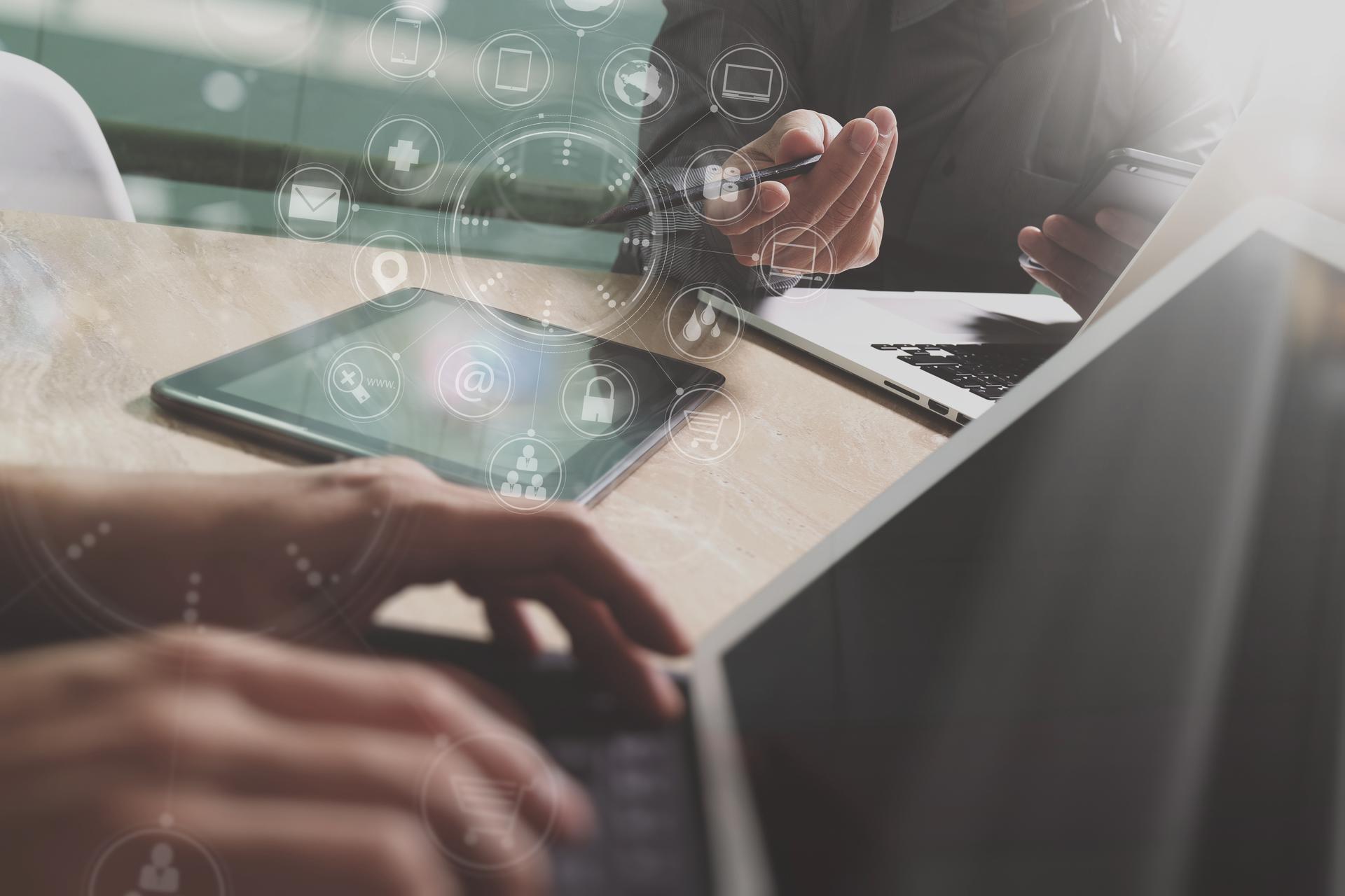 Hands typing on a laptop with a tablet nearby and a light flare in the background.