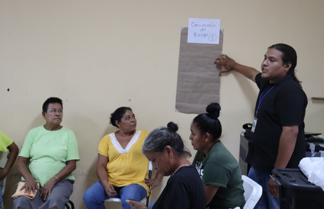 A group of people sitting in a room, with one man presenting a sheet of paper.