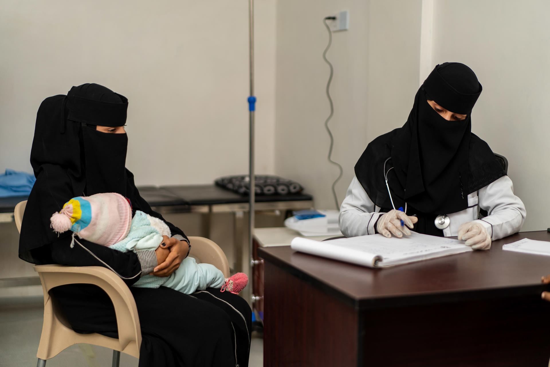 A woman in traditional attire holds a baby while another woman examines records at a desk.