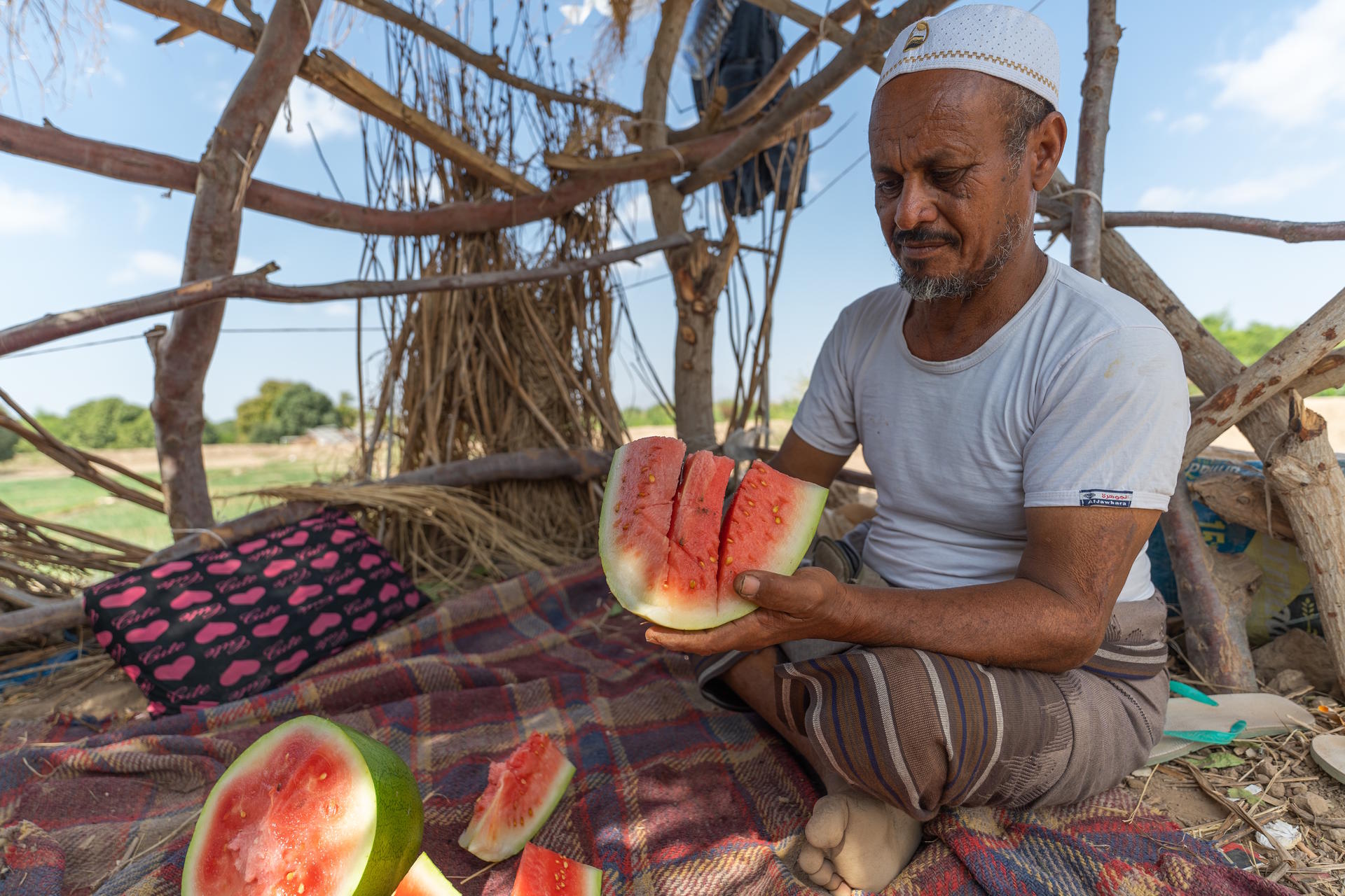 A man sitting on a mat, holding a slice of watermelon, surrounded by rustic wooden structure.