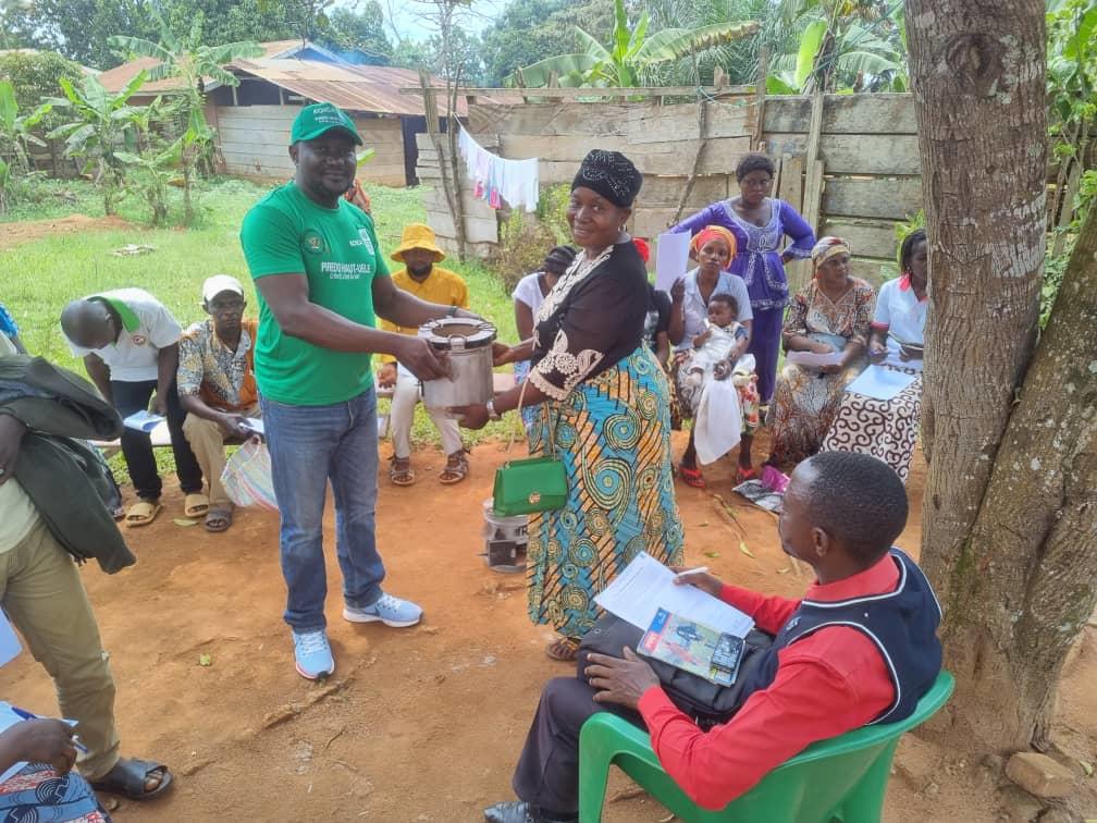 A woman in a colorful dress receives a pot from a man in a green shirt, surrounded by people seated outdoors.