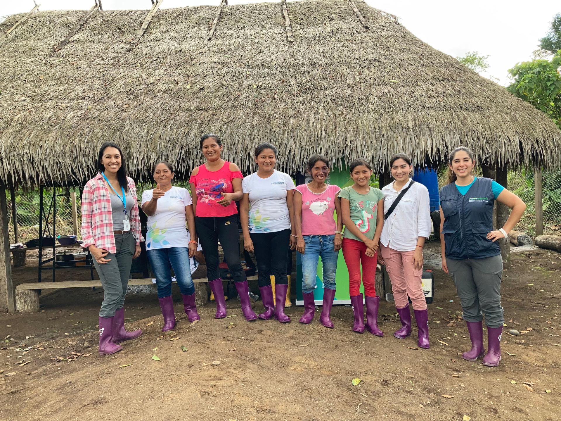 Group of eight women in rubber boots standing outside a thatched-roof hut.