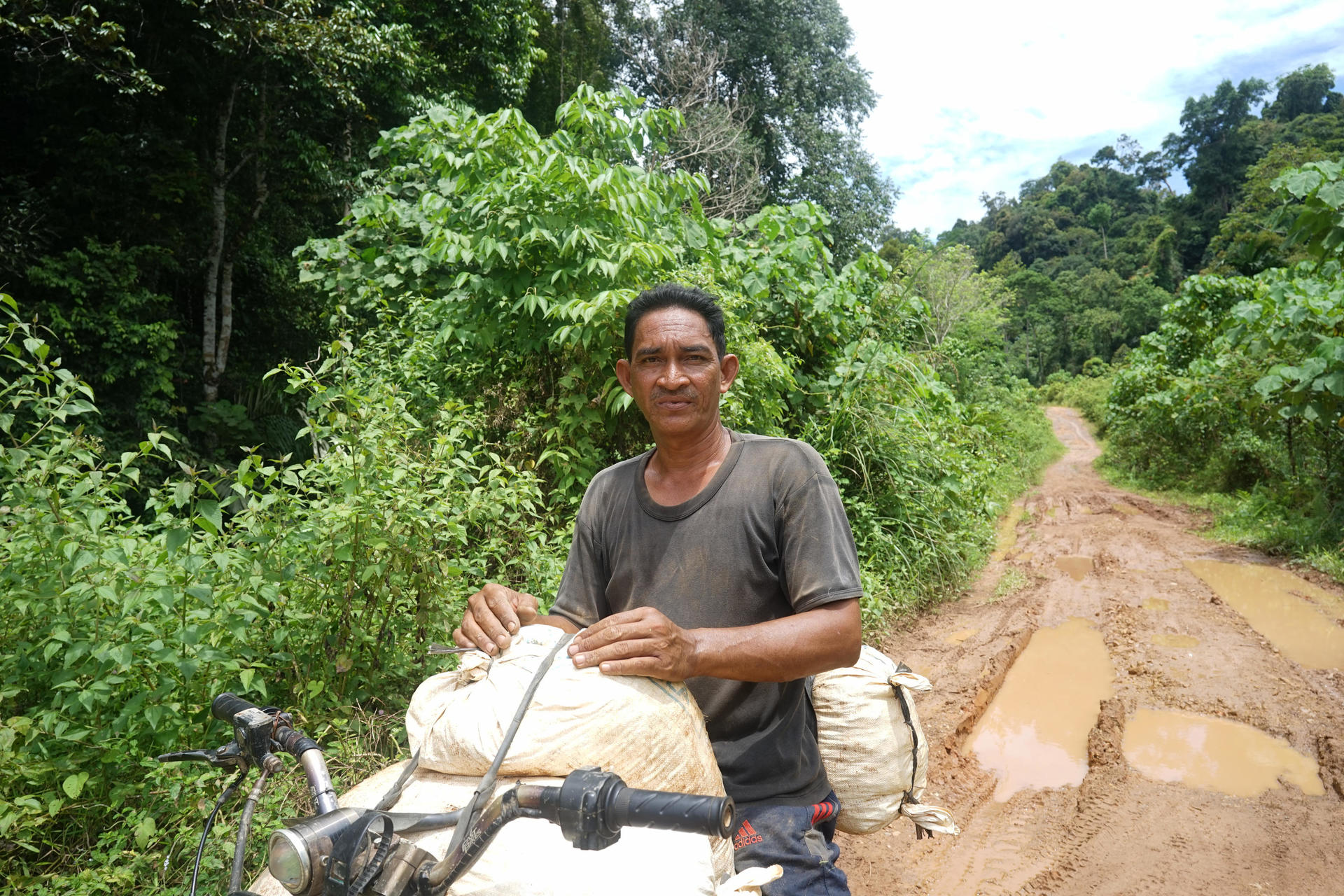 A man stands beside a bicycle on a muddy road, surrounded by lush greenery.