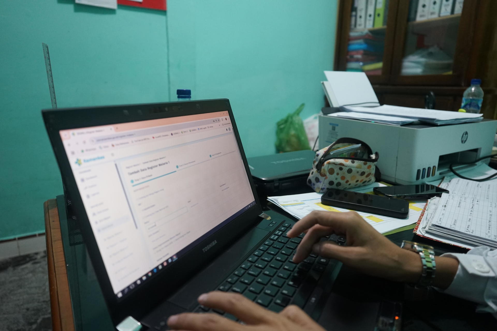 A person typing on a laptop in an office setting with documents and a printer nearby.