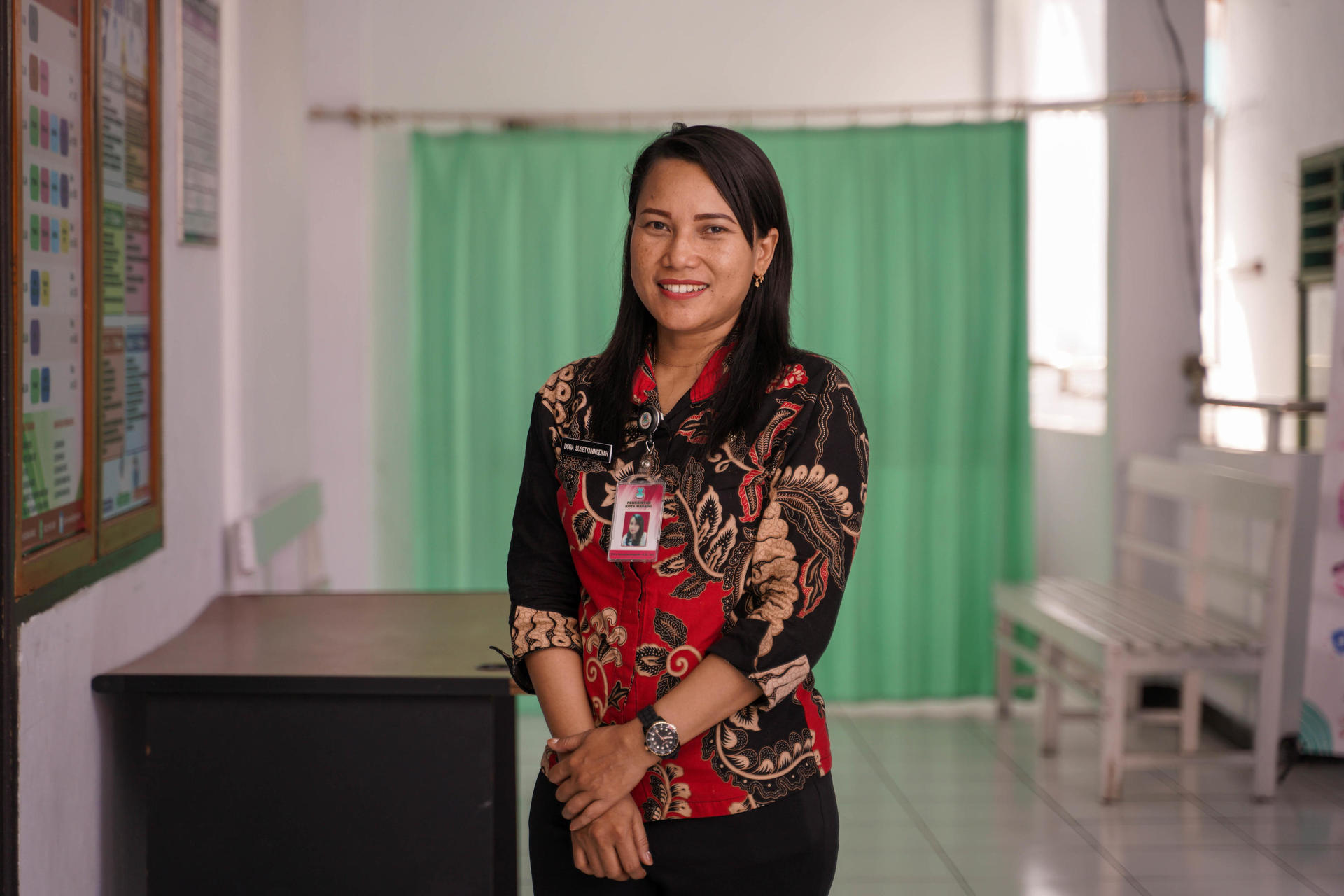 Woman smiling in a classroom with a green curtain and educational materials on the wall.