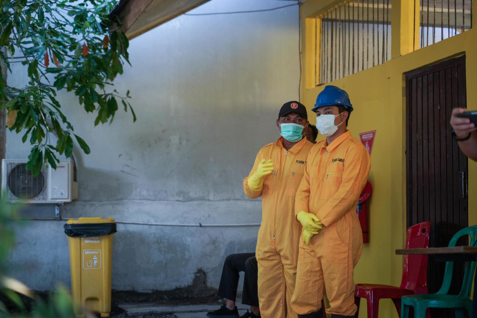 Two workers in yellow jumpsuits and hard hats stand in a sheltered outdoor area.