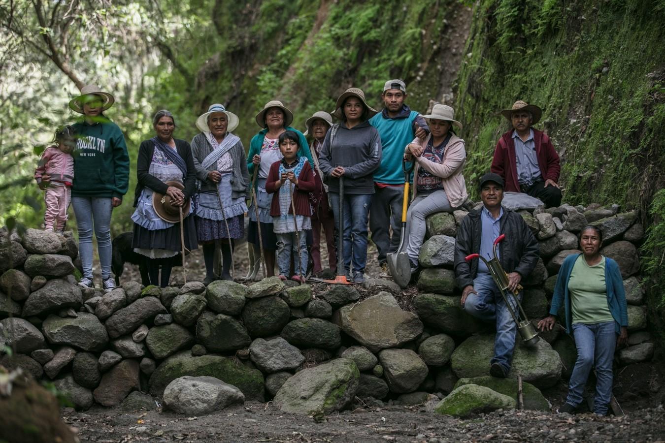 A diverse group of people stands on a stone wall in a lush, green environment.