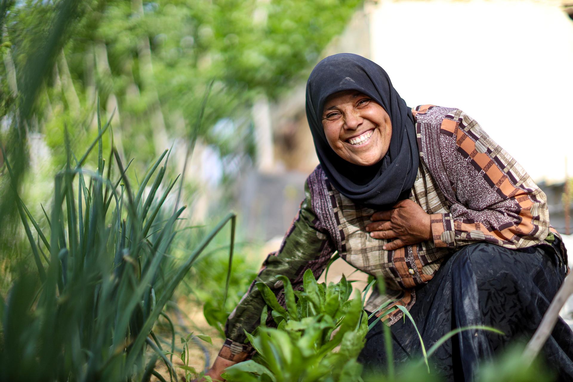 Smiling woman in a garden, surrounded by greens and plants, wearing a black hijab.