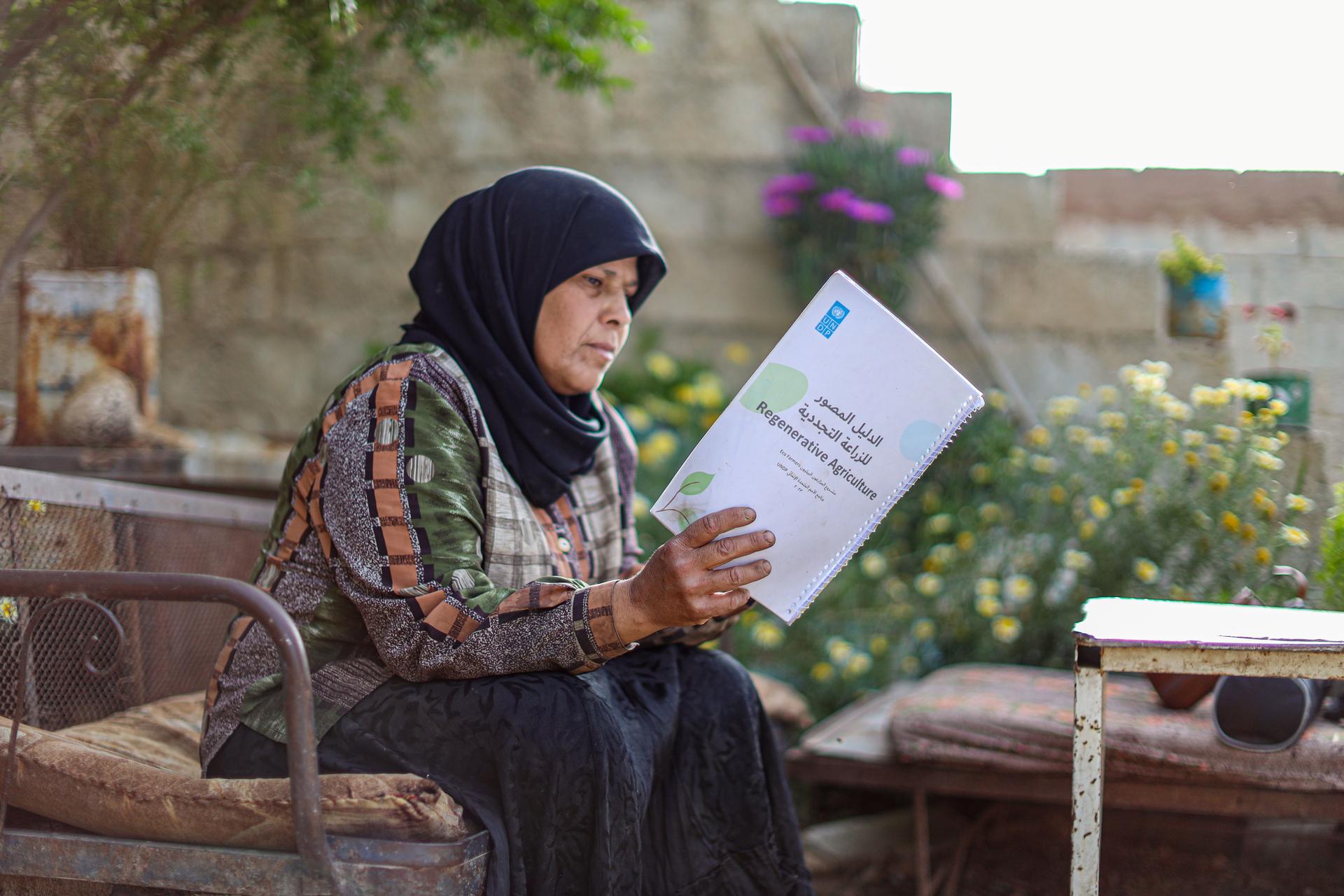 Woman in a hijab, seated outdoors, reading a booklet amidst flowers and greenery.