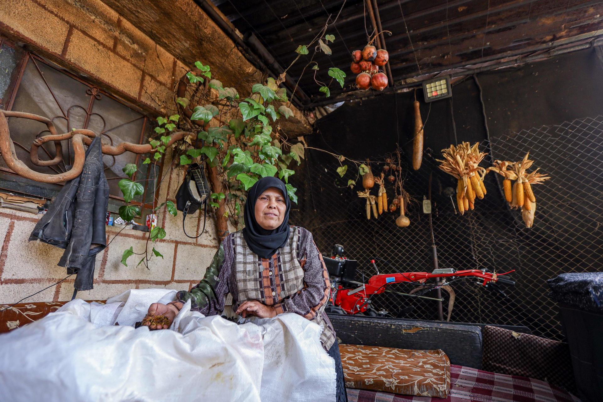 A woman in a hijab sits at a rustic table surrounded by hanging vegetables and plants.